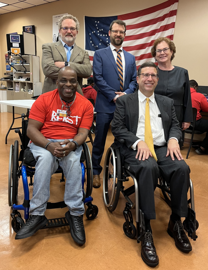 Image description: A group of people smiling for a photo, with an American flag, its stars shaped into a disability access symbol, in the background. On the back row, from left to right, Andrew Coffman, a Caucasian man with curly blond hair and a suit and tie; James Radford, a Caucasian man with a blue jacket and orange tie; and Georgia Lord, an older Caucasian woman with curly red hair. All three are wearing glasses. In the front row, from left to right, James Turner, an African-American man with a bald held and a red t-shirt in a wheelchair, and James Curtis, a Caucasian man in a dark suit and yellow tie in a wheelchair. Photo date: May 9, 2024.