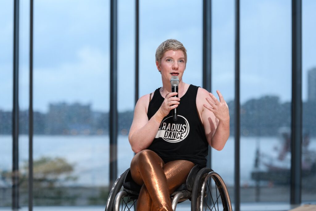 Image Description: Laurel Lawson, a pale white woman with cropped blonde hair and bright performance makeup speaks into a microphone. She is wearing a black tank top with white logo above brilliant copper leggings. The background shows the Hudson River through rain spattered glass. Photo credit: https://www.instagram.com/filipwolak/